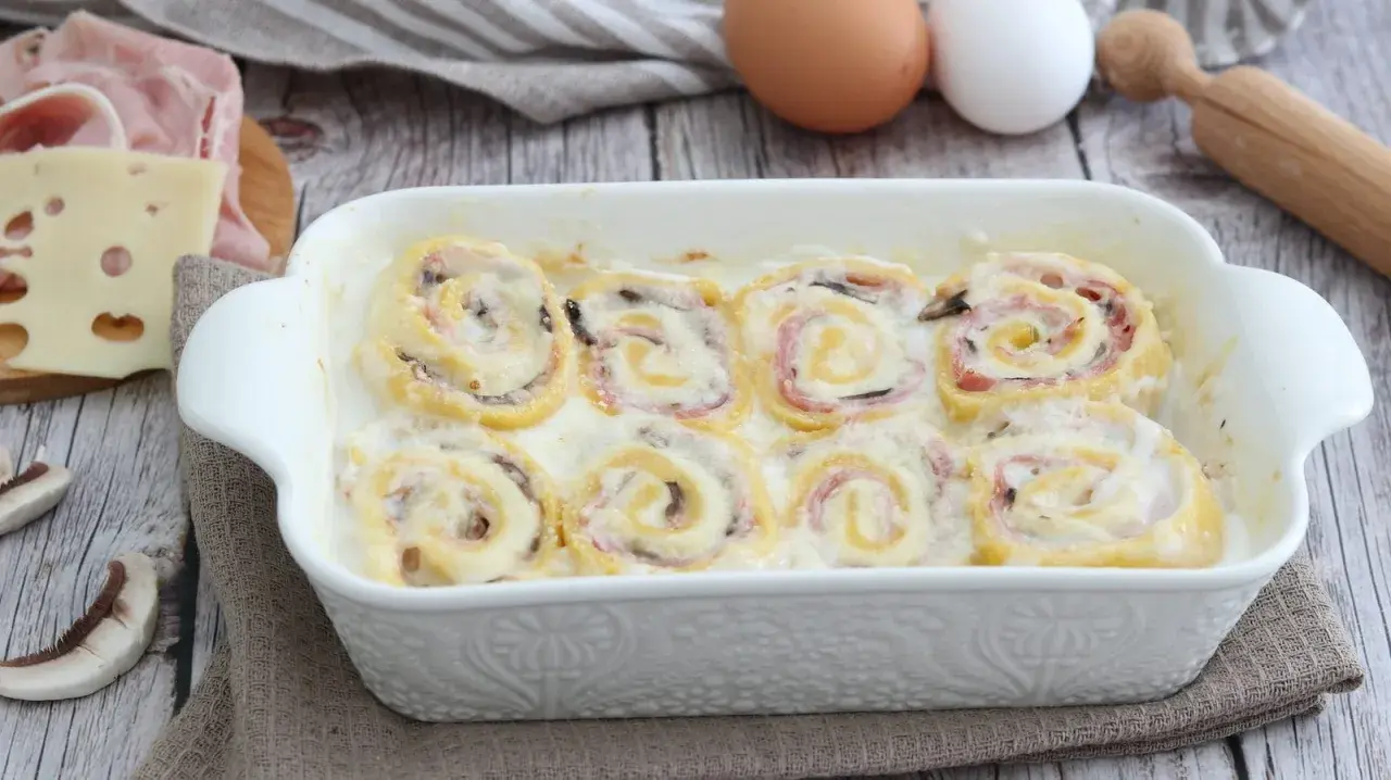 Rotolini di pasta con prosciutto e formaggio, cotti in una cremosa besciamella, sembrano piccoli nidi di rondine pronti per essere gustati.