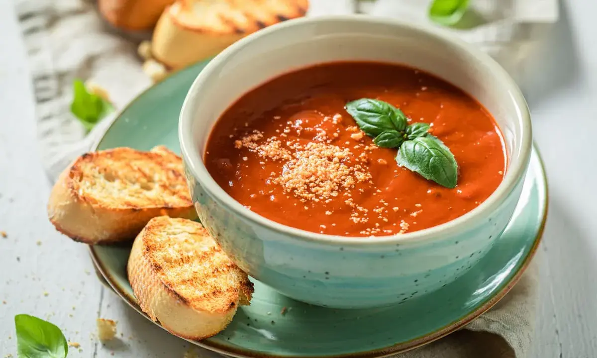 Una ciotola di cremosa zuppa di pomodoro, guarnita con basilico fresco e parmigiano, servita con crostini tostati.