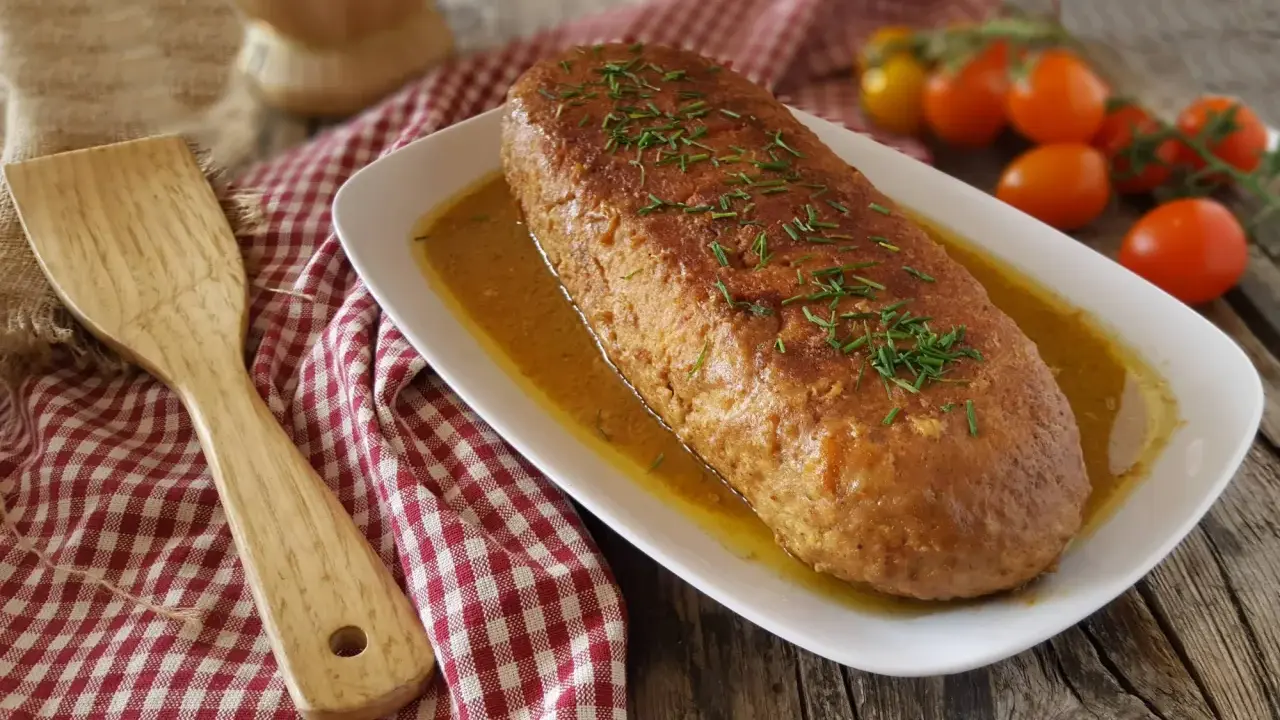 Polpettone in padella dorato e succulento, guarnito con erba cipollina, servito su un piatto bianco con salsa. Accanto, una spatola in legno e pomodorini.