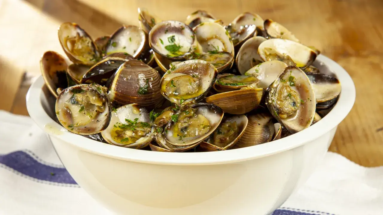 Un invitante sautè di vongole, condito con prezzemolo fresco e olio d'oliva, servito in una ciotola bianca su un tavolo di legno.