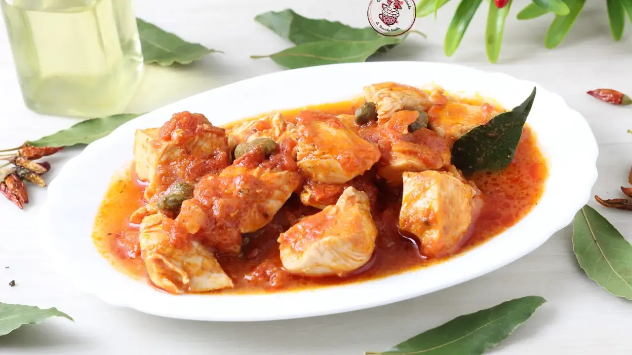 Succulento pollo alla pizzaiola in un piatto bianco, con capperi, pomodoro e foglie di alloro. Accanto, peperoncini secchi e una bottiglia d'olio.