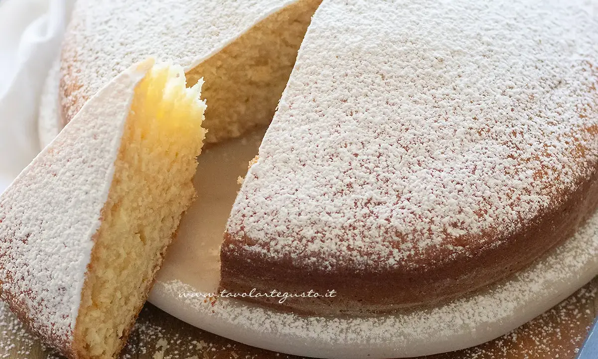 Una soffice torta senza uova, cosparsa di zucchero a velo, con una fetta tagliata che ne mostra l'interno chiaro e profumato.