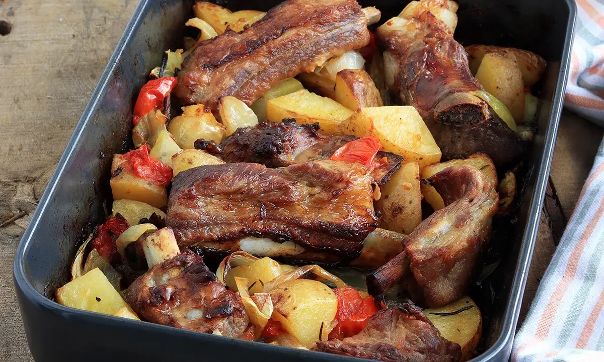 Succulente costine al forno con patate, peperoni e cipolle in una teglia scura.