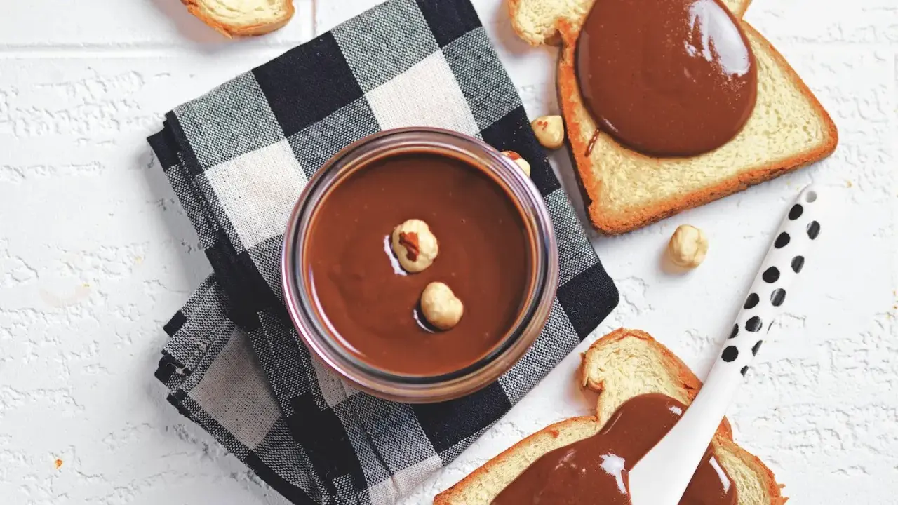Un vasetto di crema di nocciole fatta in casa, con nocciole intere, su pane tostato e tovagliolo a scacchi.