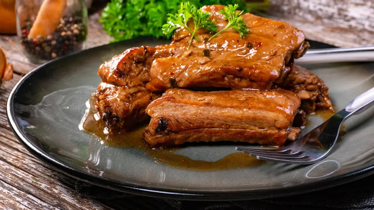Succulente costine di maiale glassate, guarnite con prezzemolo fresco, servite su un piatto scuro con una forchetta.