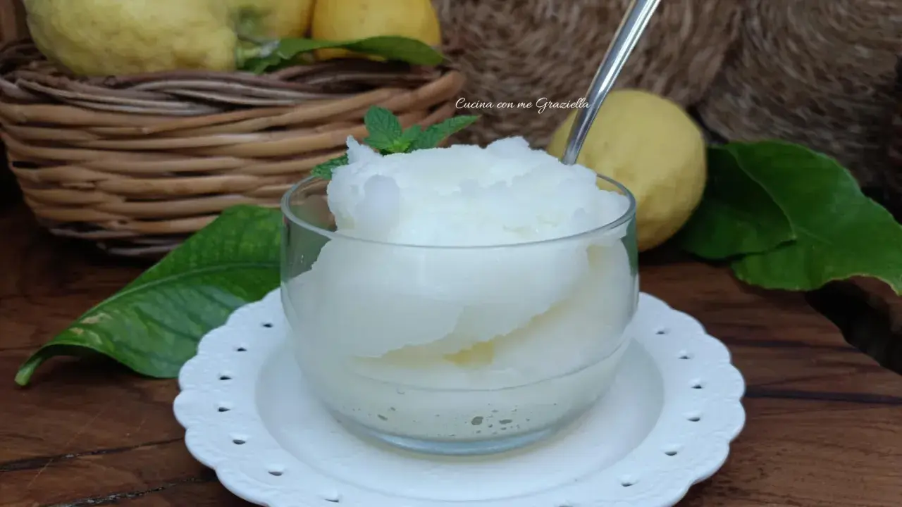 Granita al limone fatta in casa, fresca e dissetante, servita in una coppetta con foglioline di menta. Limoni freschi in un cesto sullo sfondo.