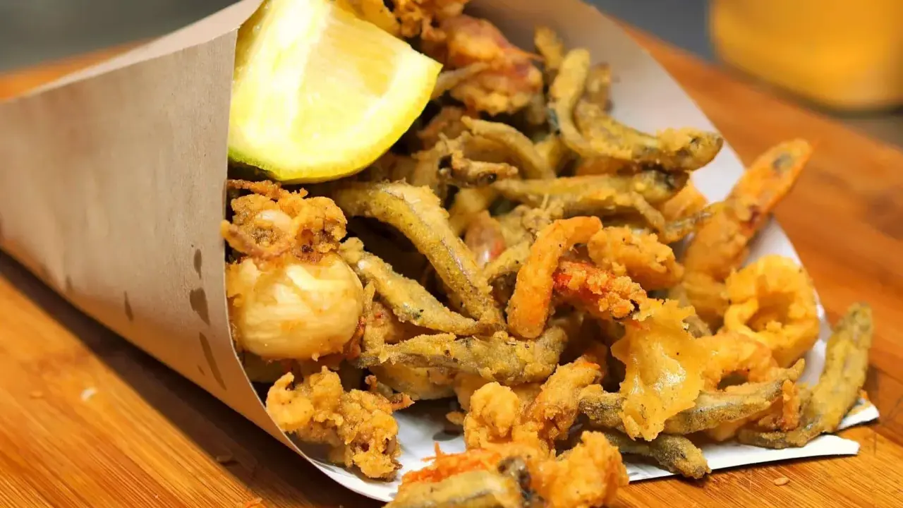 Un cono di carta marrone traboccante di una deliziosa frittura di pesce croccante, con calamari, gamberetti e pesciolini, guarnito con uno spicchio di limone.