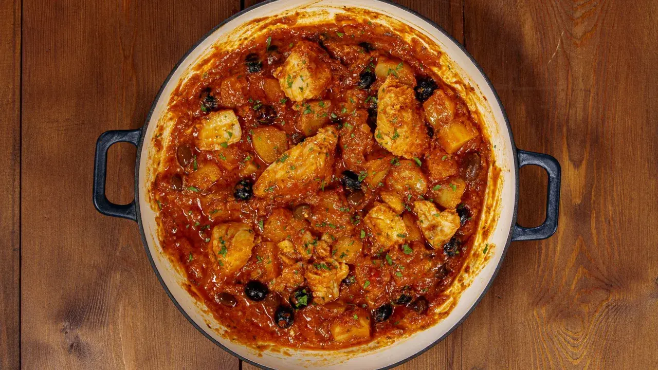 Un saporito baccalà in umido con patate e olive nere, guarnito con prezzemolo fresco, in una casseruola su un tavolo di legno.