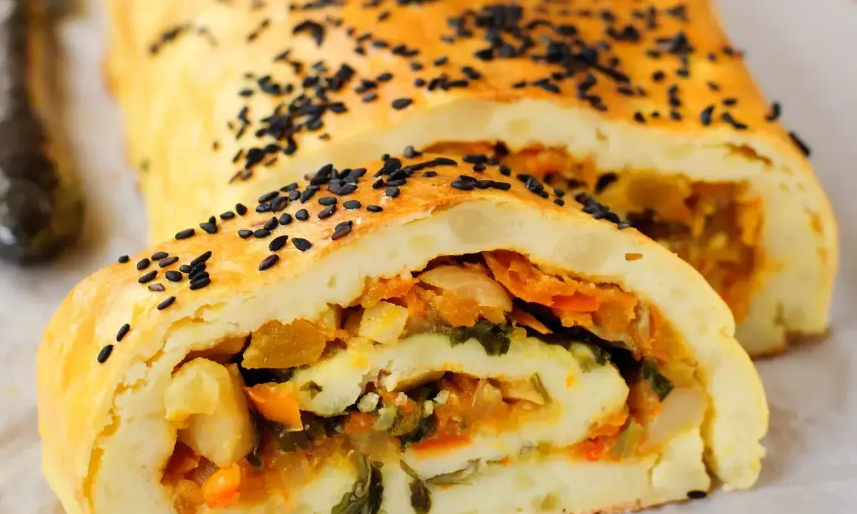 Rotolo di patate dorato, cosparso di semi neri, con un ripieno colorato di verdure.