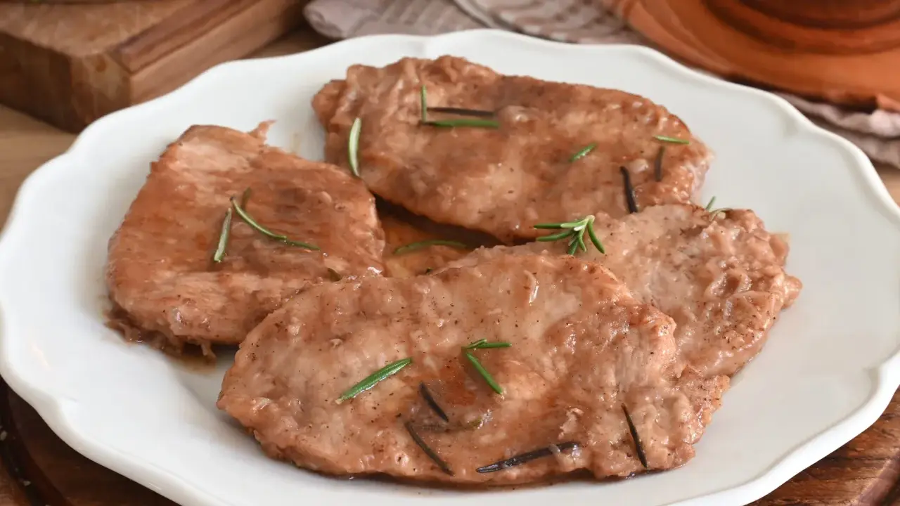 Quattro scaloppine di maiale succulente, guarnite con rosmarino fresco, servite su un piatto bianco.