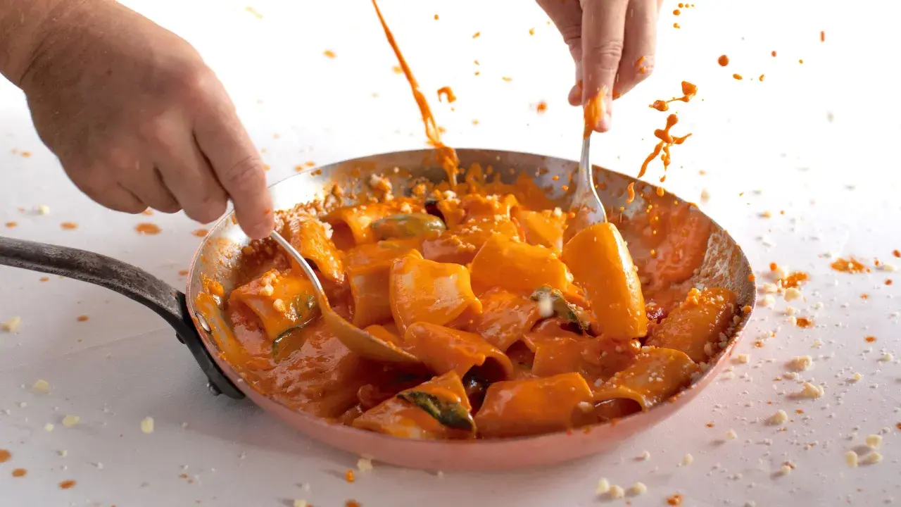 Mani che mescolano paccheri alla Vittorio in padella, con salsa arancione e schizzi di sugo.