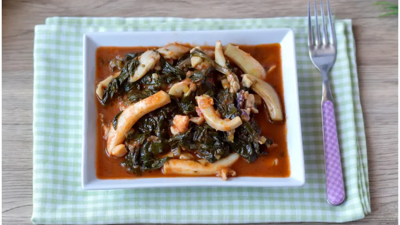Un saporito piatto di seppie in zimino, con verdure scure e salsa rossa, servito su un tovagliolo a quadretti verdi.