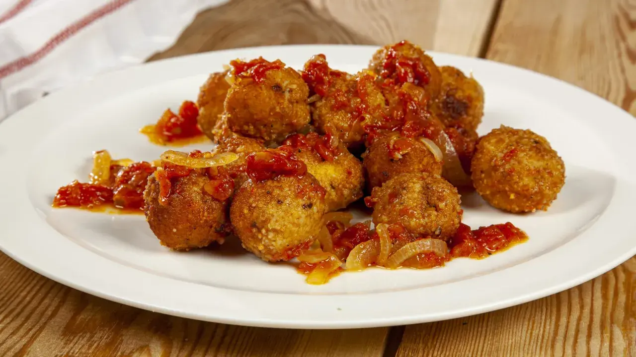 Polpette di pesce dorate e saporite, condite con un sugo rosso di pomodoro e cipolla, servite su un piatto bianco.