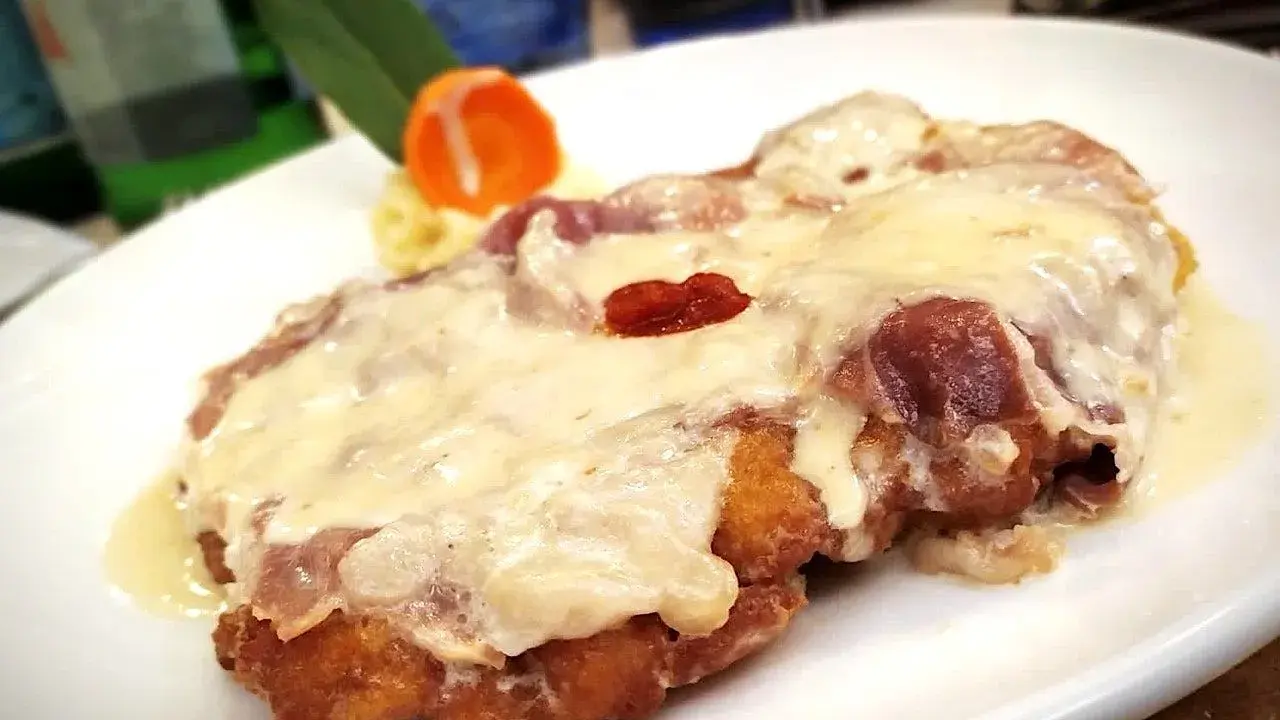 Cotoletta alla bolognese dorata e filante, con prosciutto e formaggio fuso, servita su un piatto bianco con contorno di purè e carota.