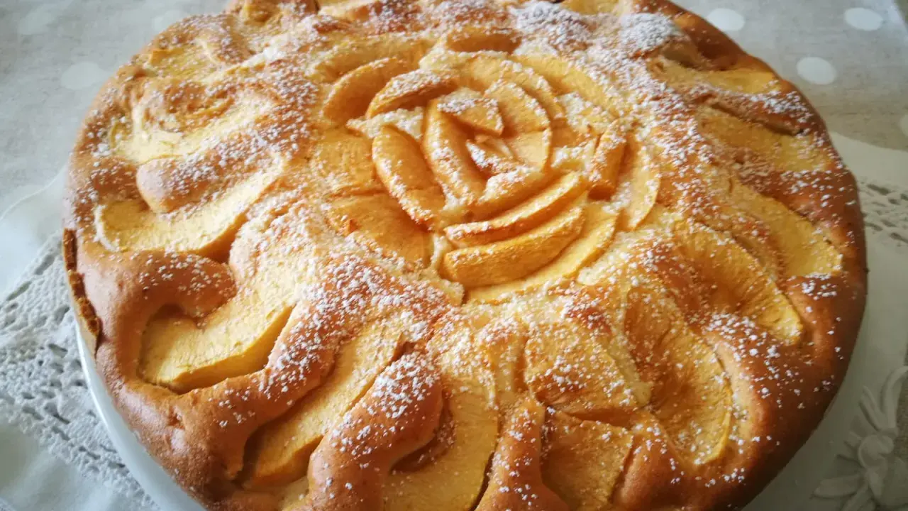 Deliziosa torta di mele senza glutine, decorata con fettine di mela disposte a rosa al centro e petali tutt'intorno, spolverata di zucchero a velo.