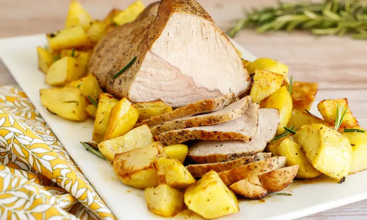 Un succulento arrosto di vitello affettato, servito su un piatto bianco con patate arrosto dorate e rametti di rosmarino.