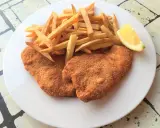 Due cotolette di pollo dorate e croccanti, accompagnate da patatine fritte e uno spicchio di limone.