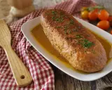 Polpettone in padella dorato e succulento, guarnito con erba cipollina, servito su un piatto bianco con salsa. Accanto, una spatola in legno e pomodorini.