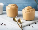 Due bicchieri di cremosa schiuma di caffè, perfetta per una ricetta crema di caffè senza panna. Due cucchiaini dorati e chicchi di caffè completano la scena.
