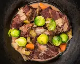 Guancia di vitello stufata con cavolini di Bruxelles, carote e cipolle in un pentolone scuro.