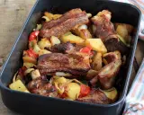 Succulente costine al forno con patate, peperoni e cipolle in una teglia scura.