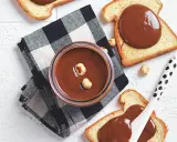 Un vasetto di crema di nocciole fatta in casa, con nocciole intere, su pane tostato e tovagliolo a scacchi.