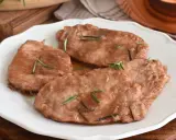 Quattro scaloppine di maiale succulente, guarnite con rosmarino fresco, servite su un piatto bianco.