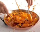 Mani che mescolano paccheri alla Vittorio in padella, con salsa arancione e schizzi di sugo.