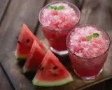 Due bicchieri di rinfrescante granita di anguria, guarniti con menta, accanto a fette di anguria fresca su un tagliere di legno.