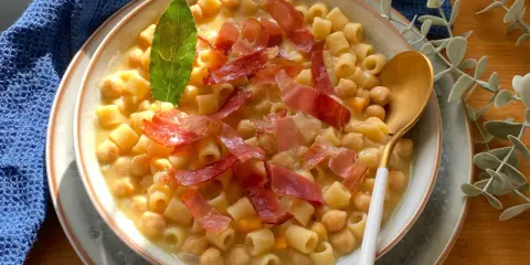 Un piatto fumante di pasta e ceci, guarnito con prosciutto crudo e una foglia di alloro.