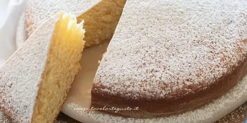 Una soffice torta senza uova, cosparsa di zucchero a velo, con una fetta tagliata che ne mostra l'interno chiaro e profumato.