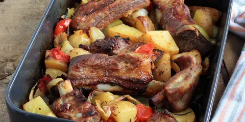Succulente costine al forno con patate, peperoni e cipolle in una teglia scura.