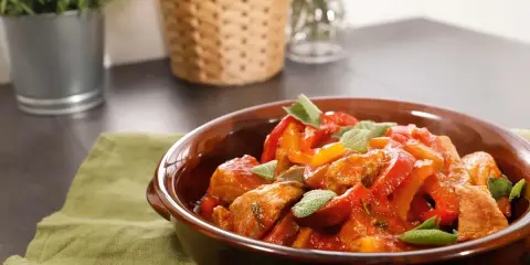 Un saporito spezzatino di vitello con peperoni rossi e gialli, guarnito con foglie di salvia fresca, servito in una ciotola di terracotta.