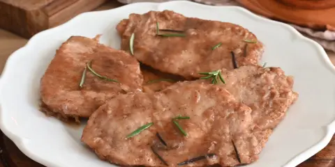 Quattro scaloppine di maiale succulente, guarnite con rosmarino fresco, servite su un piatto bianco.