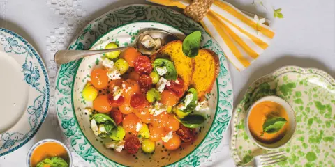 Deliziosa insalata con melone, pomodorini, formaggio e basilico, servita con pane tostato e una ciotolina di gazpacho.