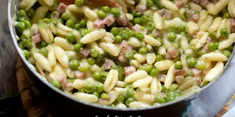 Un primo piatto invitante: pasta e piselli cremosa con cubetti di prosciutto cotto, mescolata con un cucchiaio di legno.