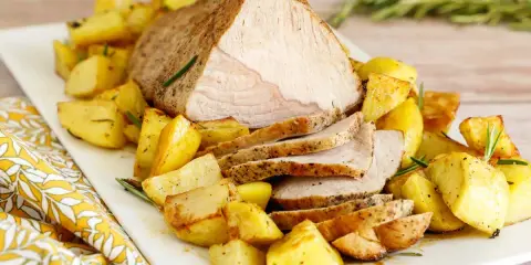 Un succulento arrosto di vitello affettato, servito su un piatto bianco con patate arrosto dorate e rametti di rosmarino.
