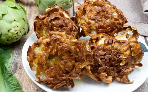 Piatto di carciofi alla giudia dorati e croccanti, con carciofi freschi sullo sfondo.