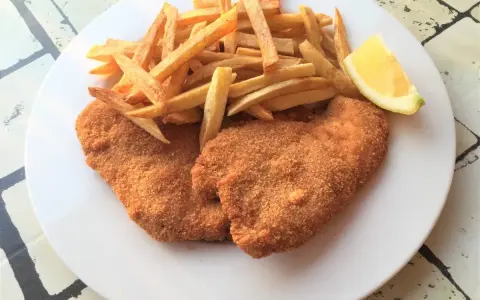 Due cotolette di pollo dorate e croccanti, accompagnate da patatine fritte e uno spicchio di limone.