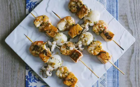 Spiedini di pesce misti, con gamberi, calamari e salmone, conditi con erbe aromatiche e serviti su un piatto bianco.