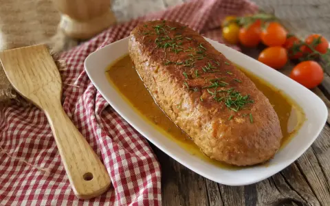 Polpettone in padella dorato e succulento, guarnito con erba cipollina, servito su un piatto bianco con salsa. Accanto, una spatola in legno e pomodorini.