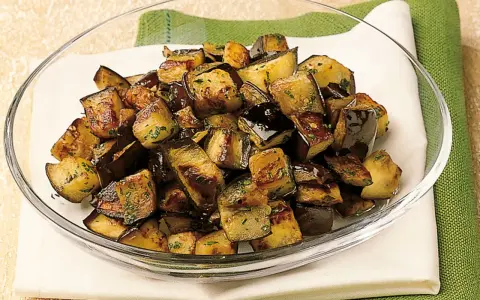 Melanzane a funghetto dorate e saporite, condite con erbe aromatiche, servite in una ciotola di vetro.
