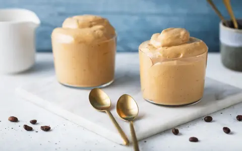 Due bicchieri di cremosa schiuma di caffè, perfetta per una ricetta crema di caffè senza panna. Due cucchiaini dorati e chicchi di caffè completano la scena.
