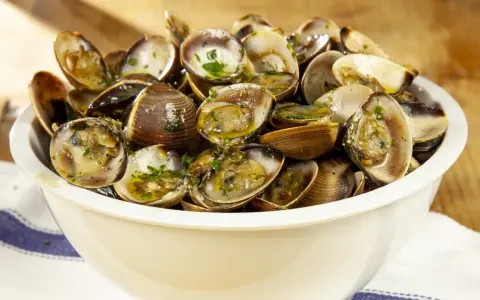 Un invitante sautè di vongole, condito con prezzemolo fresco e olio d'oliva, servito in una ciotola bianca su un tavolo di legno.