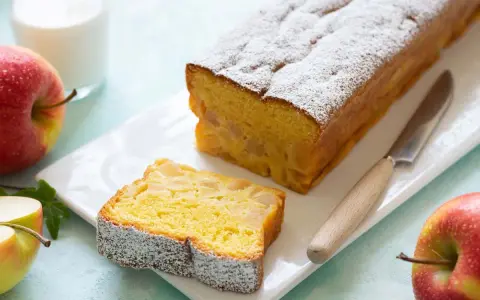Un delizioso plumcake alle mele, cosparso di zucchero a velo, con una fetta tagliata e mele rosse fresche accanto.