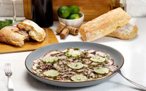 Delizioso carpaccio di polpo, guarnito con fette di lime e erbe aromatiche, pronto per essere gustato con pane fresco e vino.
