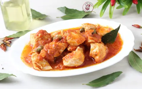 Succulento pollo alla pizzaiola in un piatto bianco, con capperi, pomodoro e foglie di alloro. Accanto, peperoncini secchi e una bottiglia d'olio.