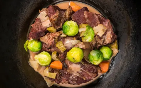 Guancia di vitello stufata con cavolini di Bruxelles, carote e cipolle in un pentolone scuro.