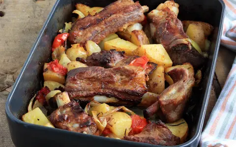 Succulente costine al forno con patate, peperoni e cipolle in una teglia scura.