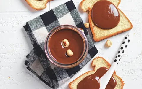Un vasetto di crema di nocciole fatta in casa, con nocciole intere, su pane tostato e tovagliolo a scacchi.